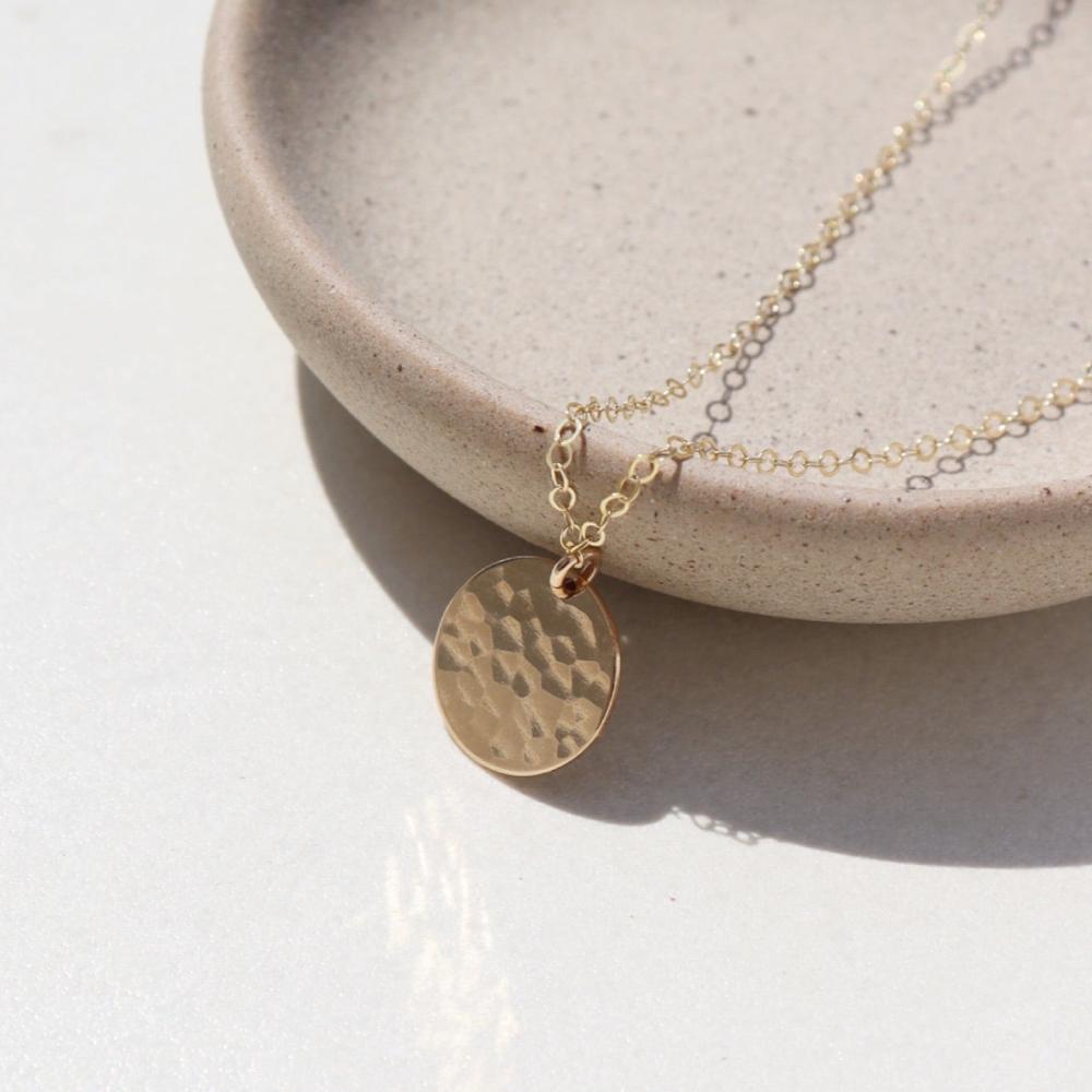 14k gold fill Moon drop necklace laid on a plate in the sunlight. This necklace features our simple chain met by a hammered coin, making it look like the moon.