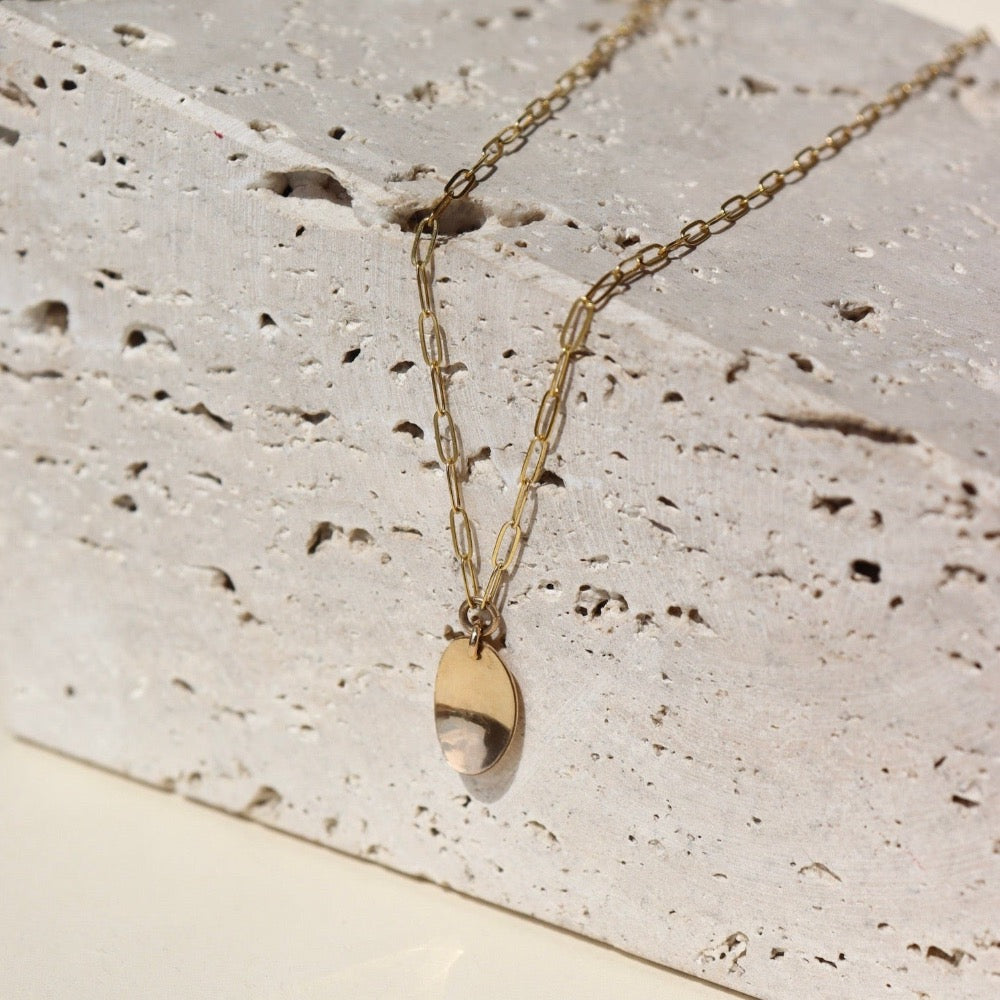 14k gold fill Olivia Necklace laid on a tan stone in the sunlight. This necklace features out rosette chain that looks like paper clips linked together as well as an oral charm that is hammered on one side