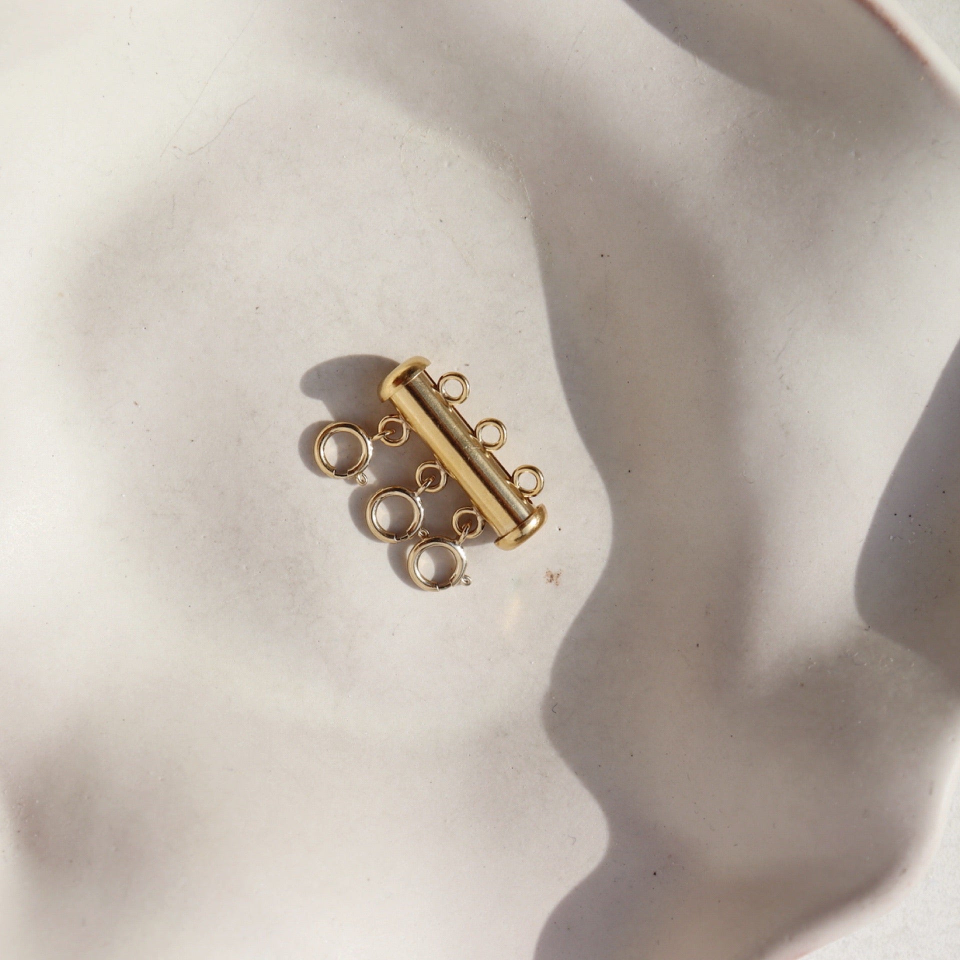 A gold necklace layering clasp with three necklace attachment points laid on a cream backdrop.