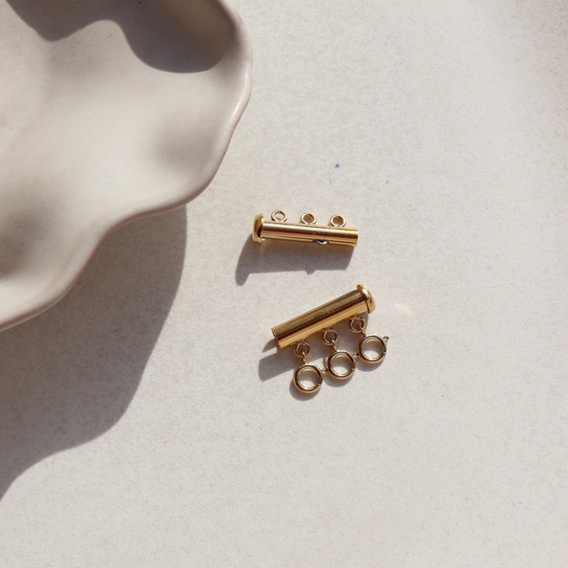 A gold necklace layering clasp with three necklace attachment points laid on a stone plate in the sunlight.