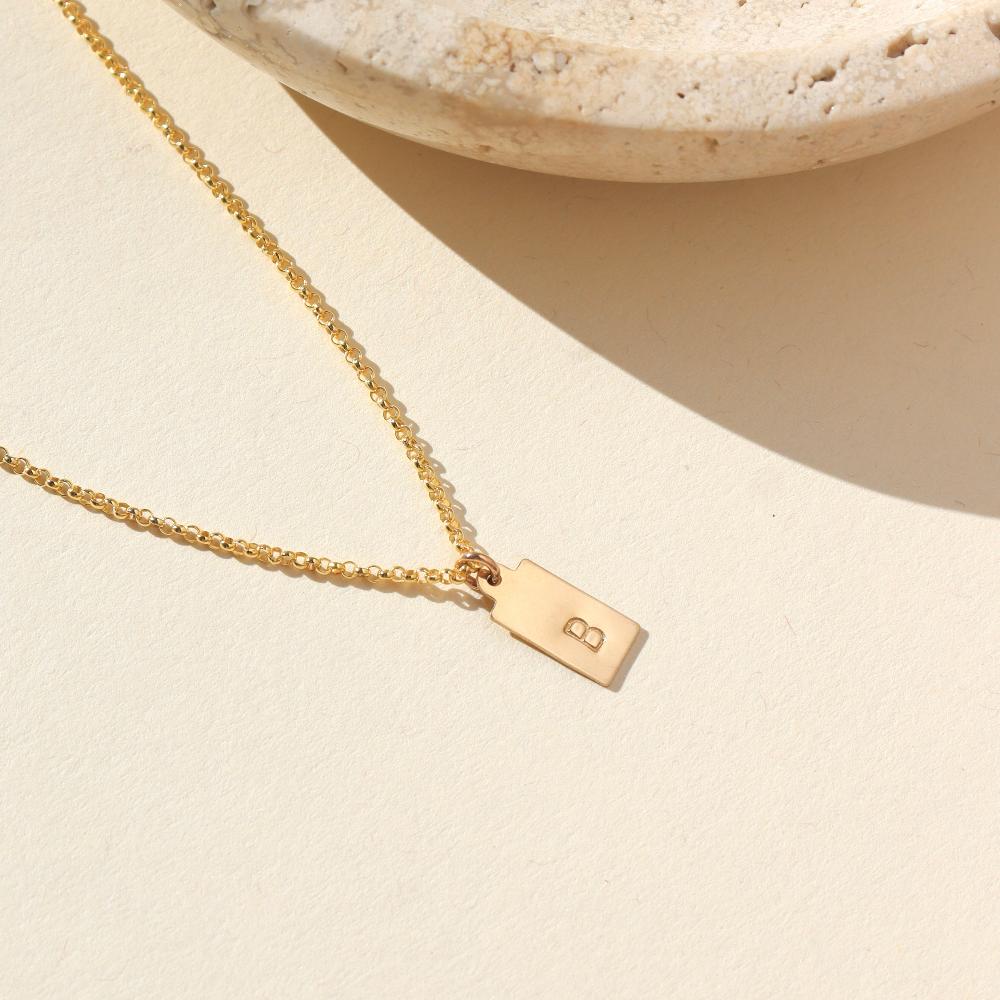 A gold rolo chain with a stamped mini tag with the letter "B". Necklace is displayed next to a stone dish.