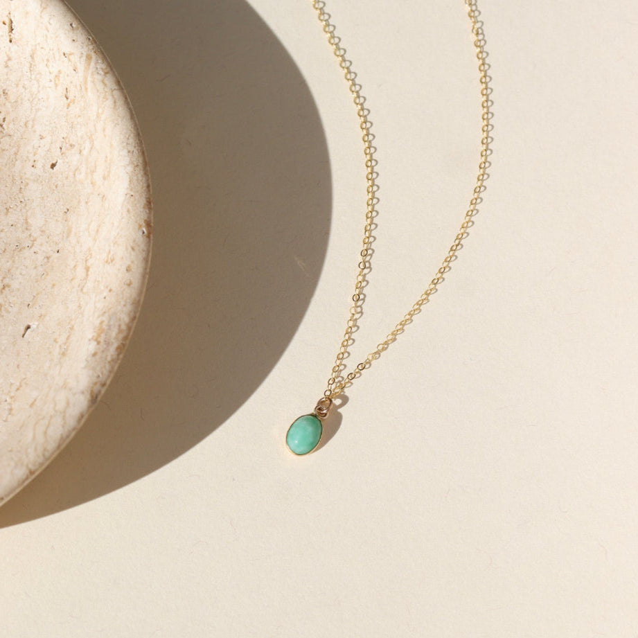 A 6x8mm Amazonite Gemstone set in a 14k gold fill bezel. The necklace features a thin standard chin. Necklace is displayed on a cream back drop in the sunlight.