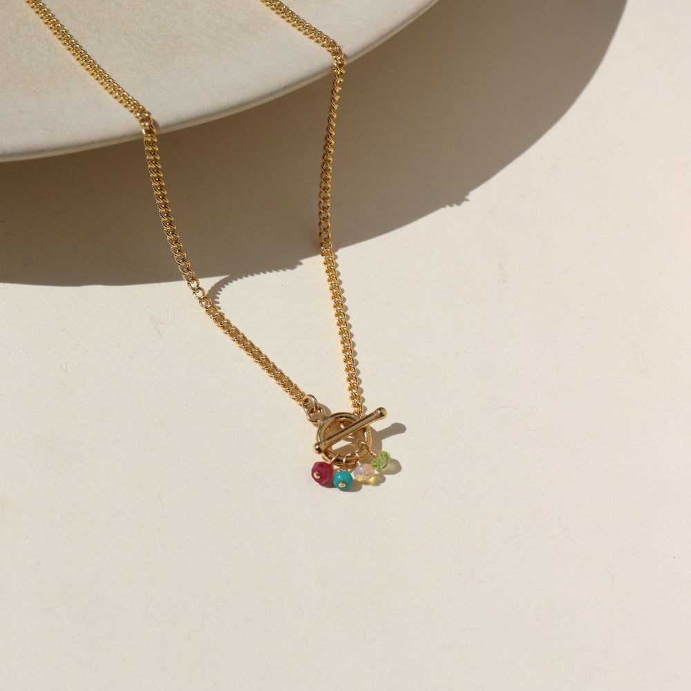 A 14k gold fill toggle necklace featuring a curb chain with four gold wire wrapped birth stones dangling from the toggle. Necklace is displayed on a cream back drop.