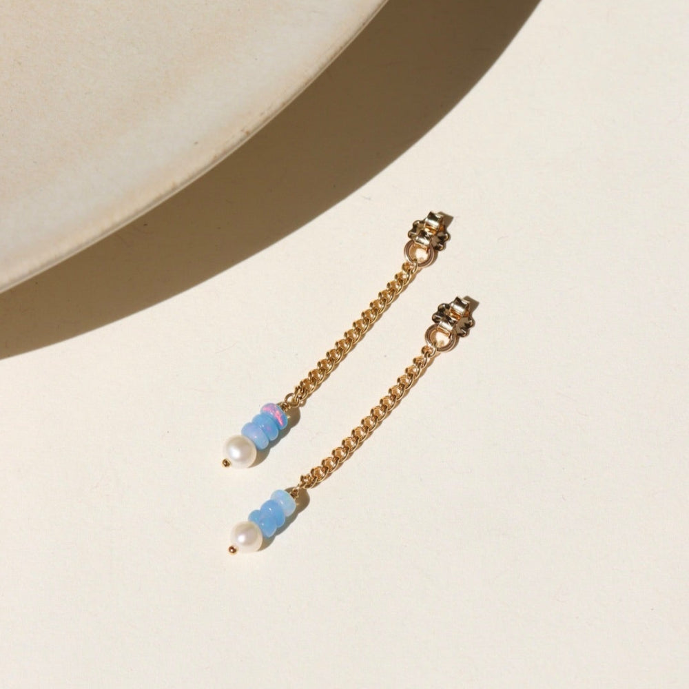 A 14k gold fill back drop with a dainty curb chain, with three opal gemstones and one mini pearl set at the bottom, the earrings are displayed on a cream backdrop in the sunlight.