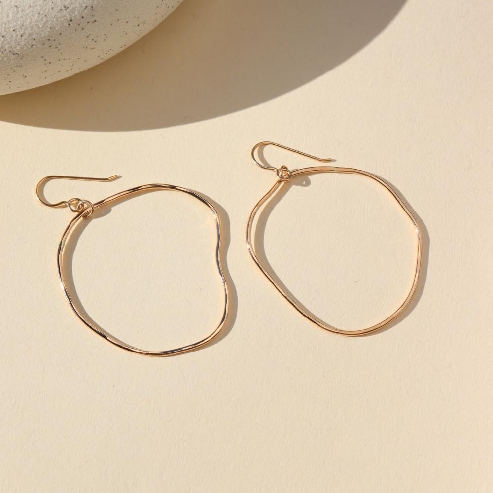 A gold large hoop earrings, each uniquely shaped, hang elegantly from simple hooks. Earrings are displayed on a cream backdrop.