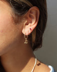 Side profile shot of a woman with her hair pulled back, showcasing a four-piercing ear stack in natural sunlight: a small gold hoop with a dangling cherry charm in her lobe, two thin gold huggie hoops in her second and third placements, and a tiny red gemstone stud.