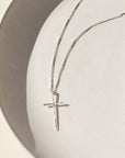 A sterling silver thin bar and link chain featuring a 1 1/2" hammered cross pendant. The necklace is displayed on a white dish.