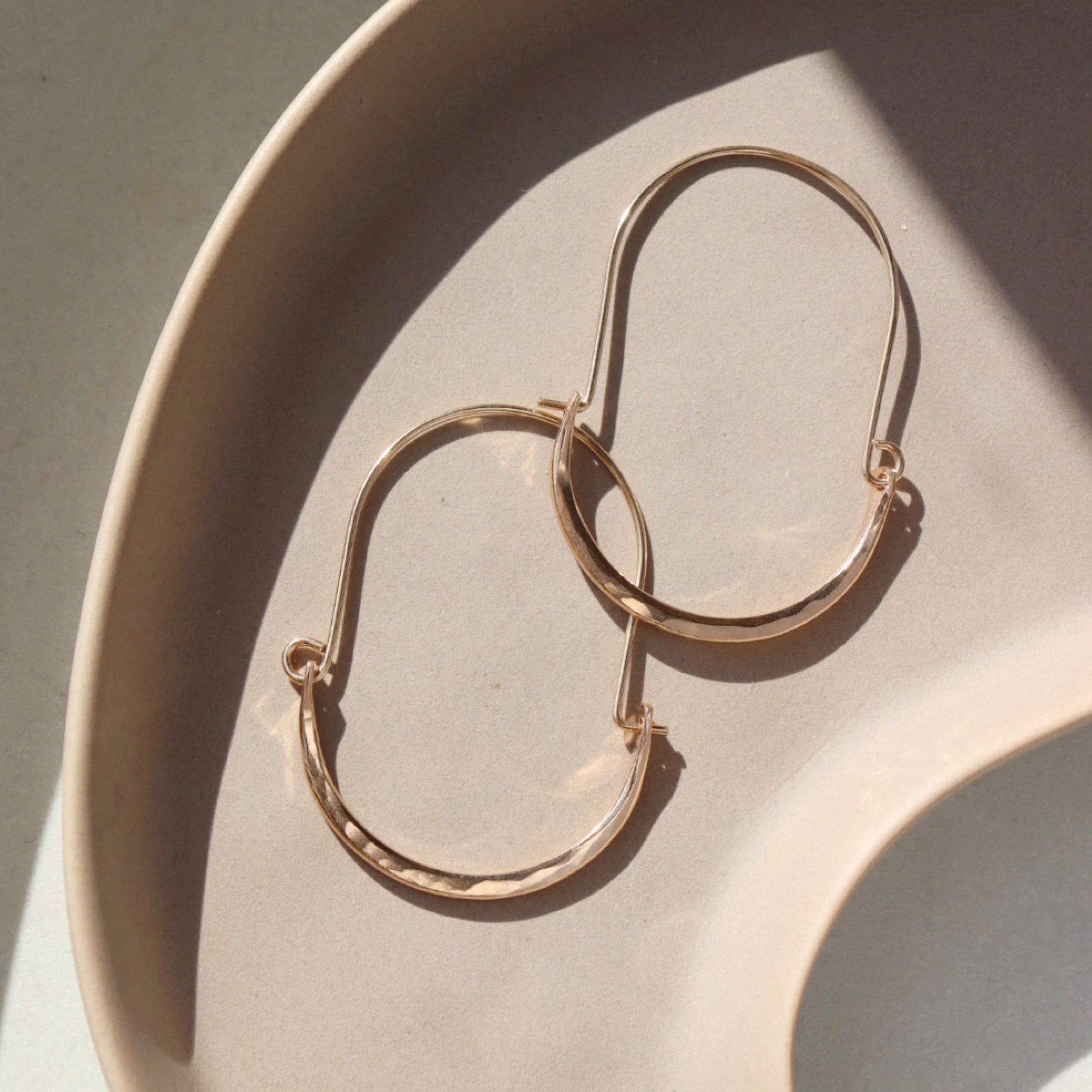14k gold fill hammered Paloma hoops, earrings are placed on a peach colored plate in the sunlight. These earrings feature a wire around 1/2 inch shaped like an oval, then followed by the other half of the oval that is hammered.
