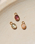 Three oval gemstone charms in pink, green, and peach tones set in gold bezels, displayed on a neutral textured surface in warm natural light.