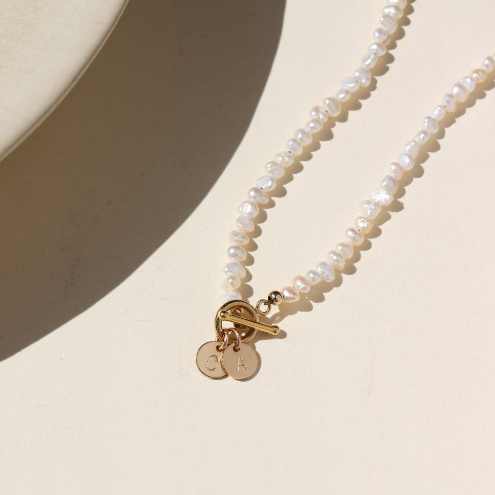 A fully beaded pearl toggle necklace with two 1/4" stamped disc with an "C" and a "A". The necklace is displayed on a cream back drop.
