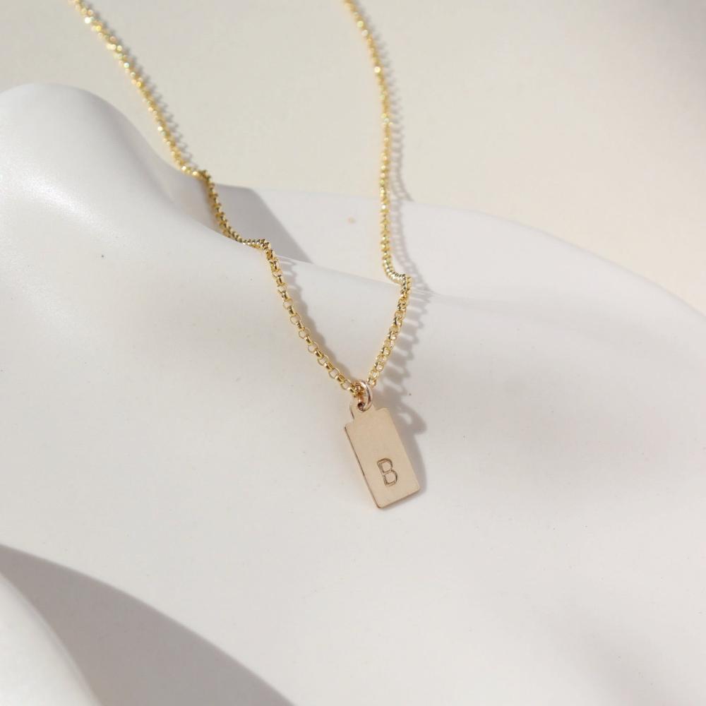 A gold rolo chain with a stamped mini tag with the letter "B". Necklace is displayed over a white dish.