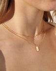 Red head model wearing the mini monogram tag necklace. This necklace features a gold rolo chain with a small stamped "B" rectangle pendant.