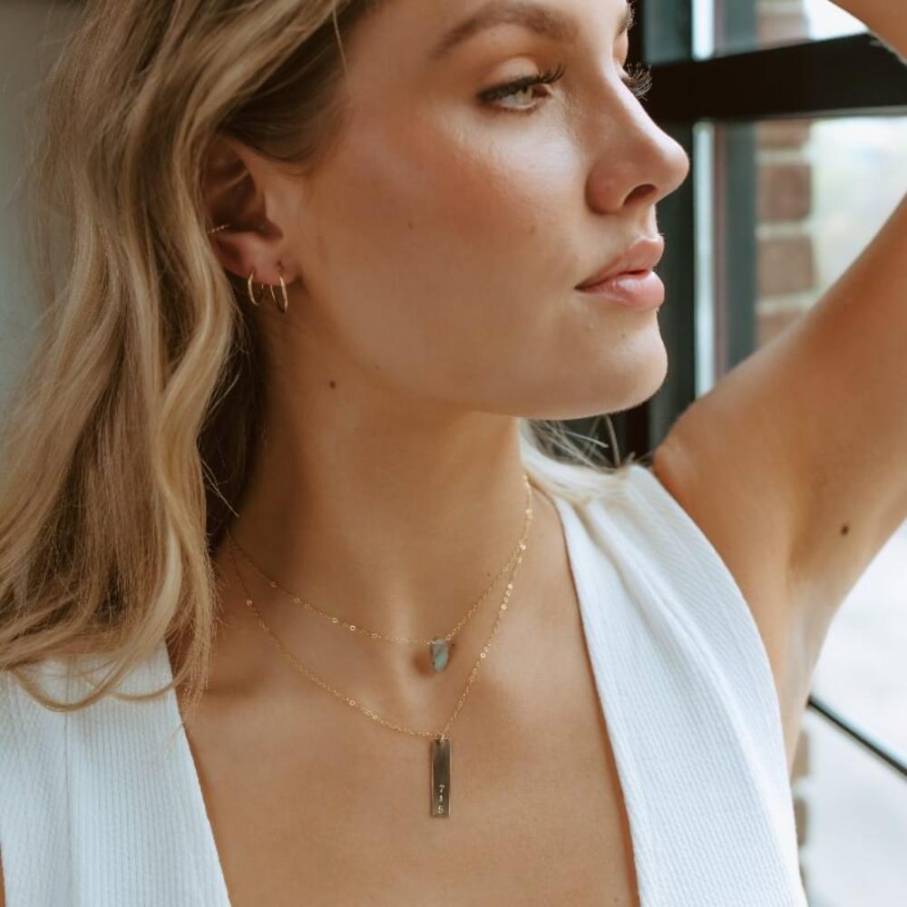 Gold hand stamped, area code necklace with delicate chain, paired with a thin labradorite gemstone necklace worn by the model.