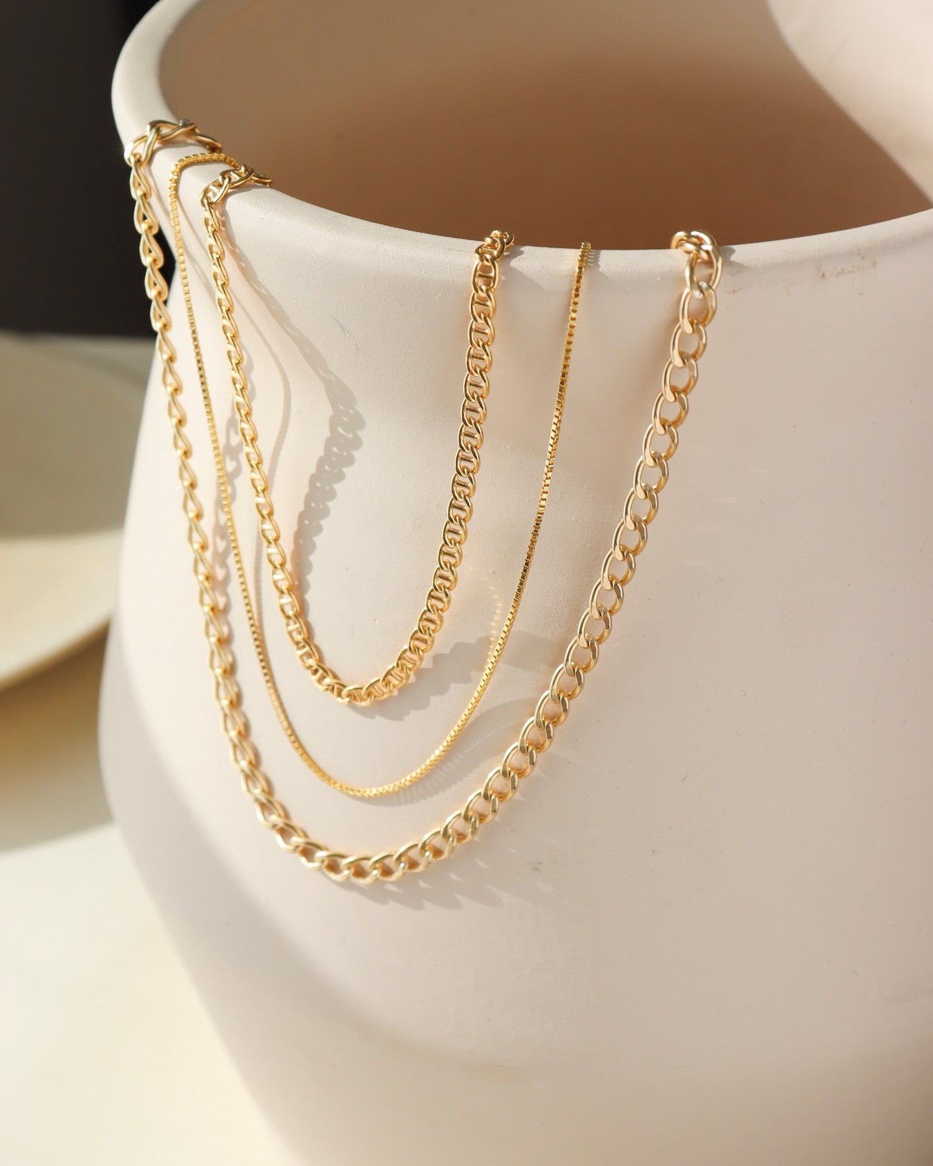A collection of gold chain necklaces in various link styles and thicknesses draped over the rim of a matte cream ceramic vase.