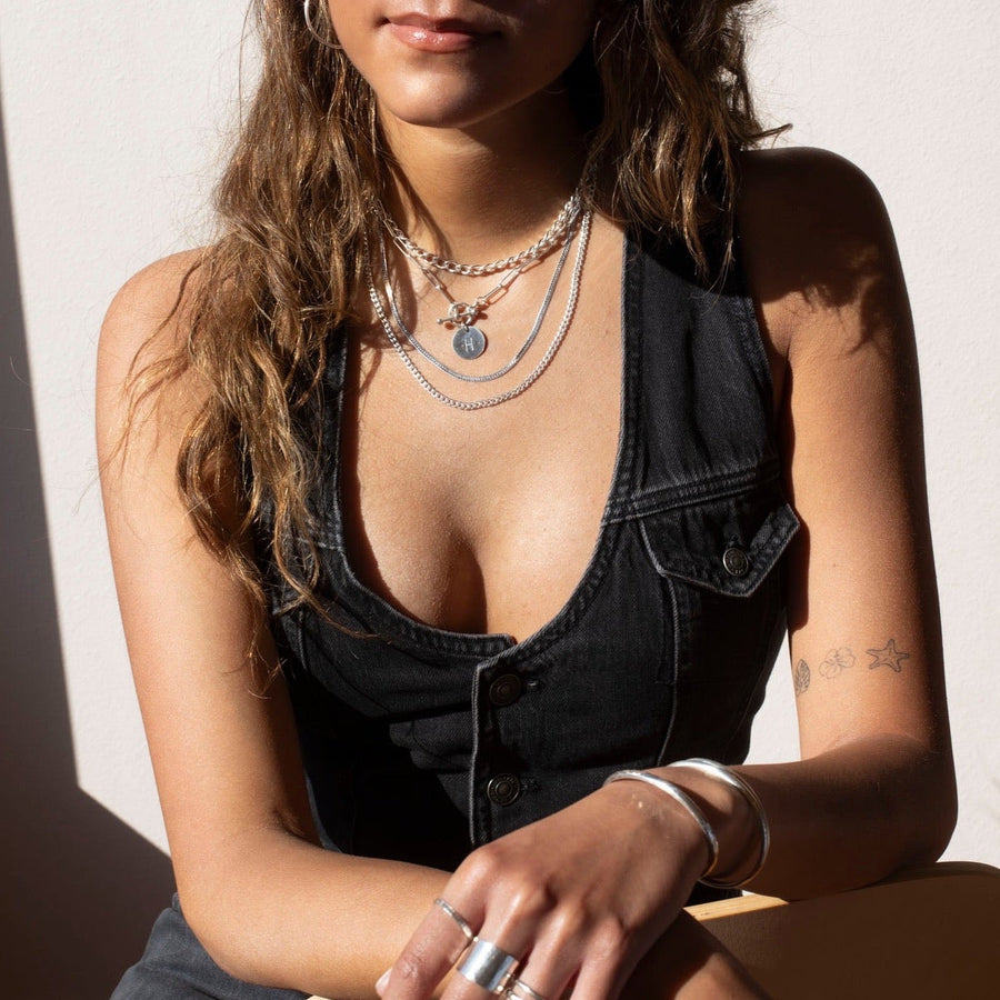 sterling silver chain link necklace with a toggle clasp and monogrammed "B" flat disk charm, photographed on a model with long brown hair, wearing silver jewelry, a black denim vest, and a black cowboy hat