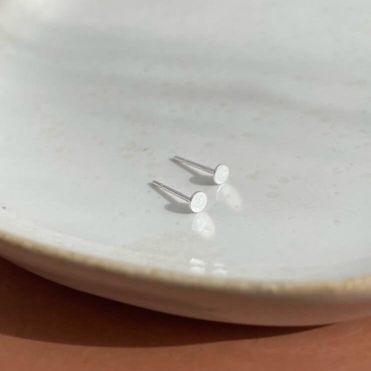 Tiny handmade sterling silver circle studs sitting on a white jewelry dish