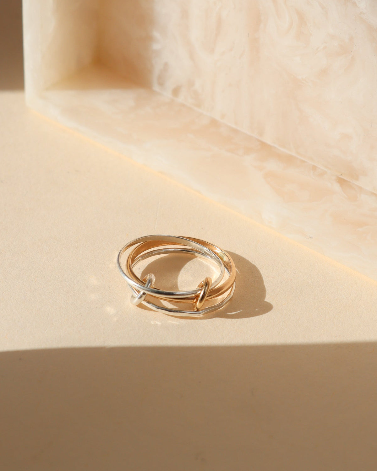 Mixed-metal ring featuring interlocking gold and silver bands with small loop accents, displayed on a cream surface in warm sunlight.