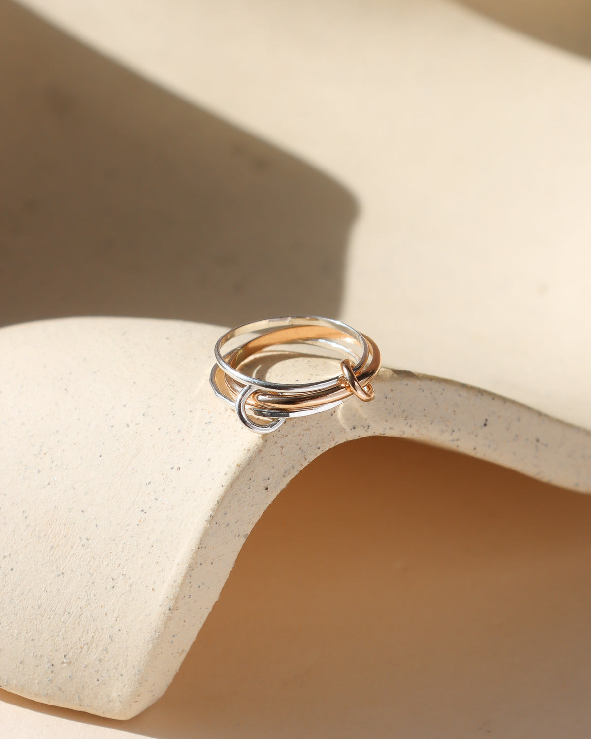 Close-up of an intertwined gold and silver mixed-metal ring resting on a textured ceramic surface in soft natural light.
