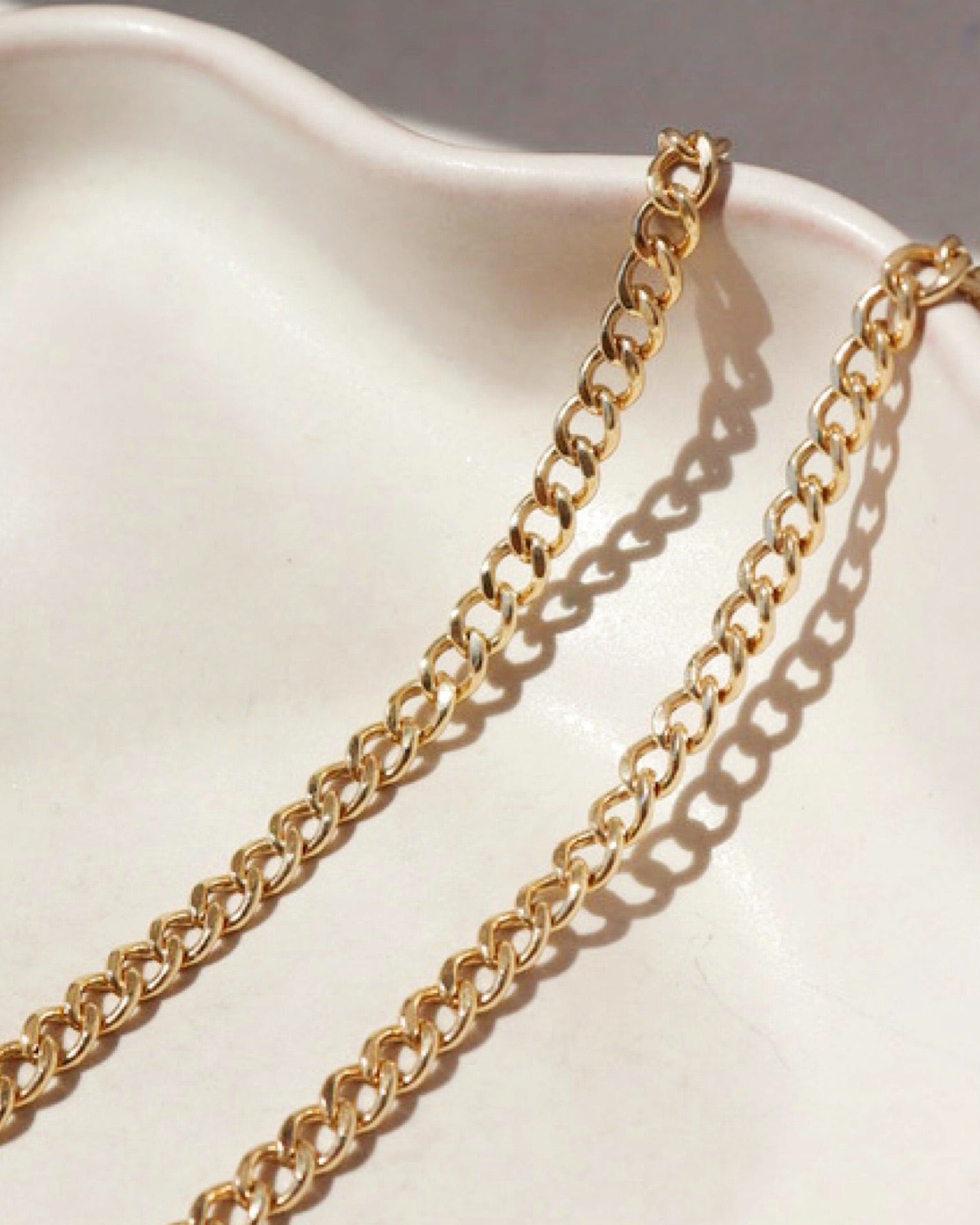 Extreme macro close-up of a gold curb chain resting on a smooth, off-white ceramic dish under direct sunlight.