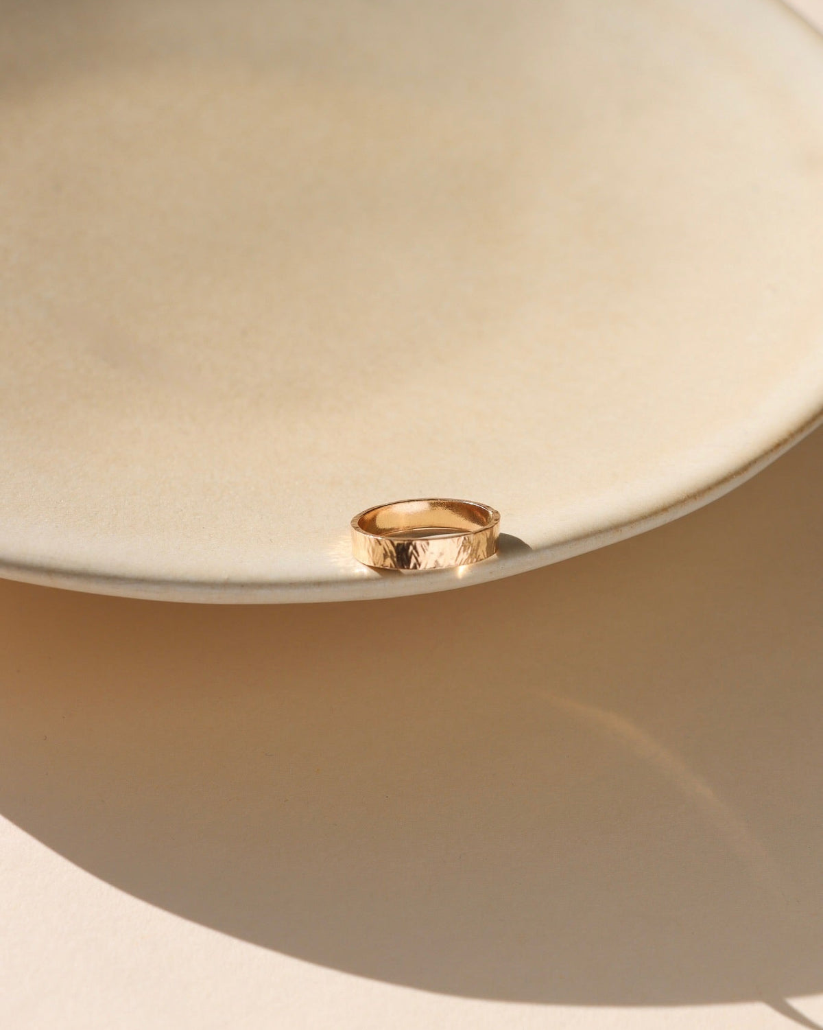 Textured gold band ring displayed on the edge of a neutral ceramic dish in warm sunlight.