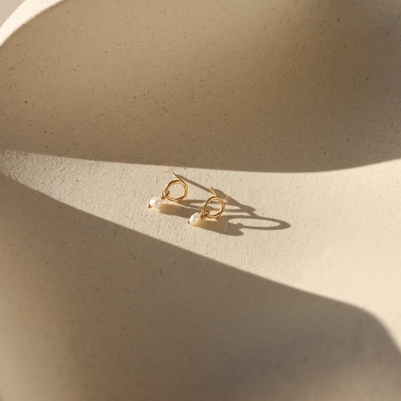 Product photo of 14k gold fill circle stud earrings featuring freshwater pearl drop accents, displayed on a textured ceramic surface in warm natural sunlight.
