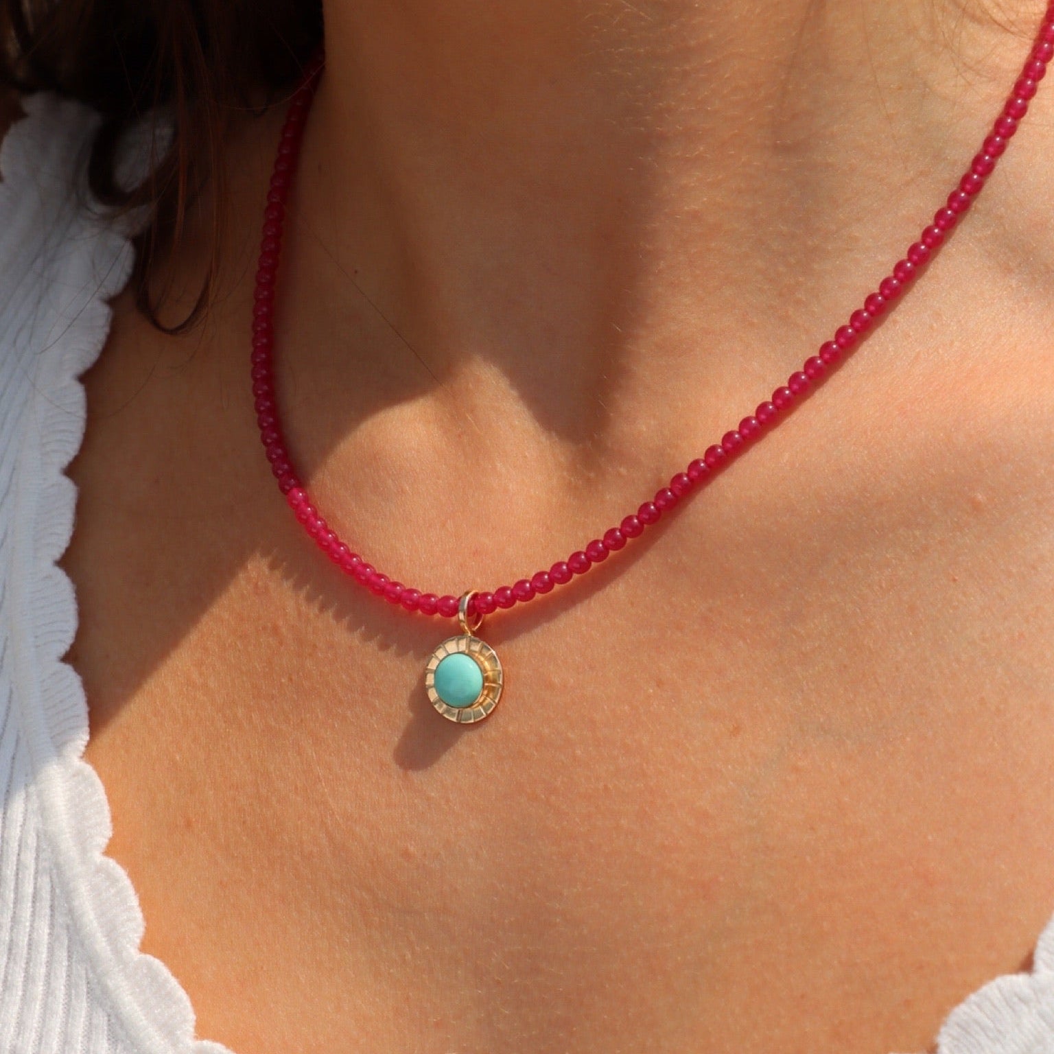 Close-up of a person wearing a bright pink beaded necklace with a small round gold pendant featuring a turquoise stone in a textured bezel, paired with a white ruffle-trim top in warm natural sunlight.