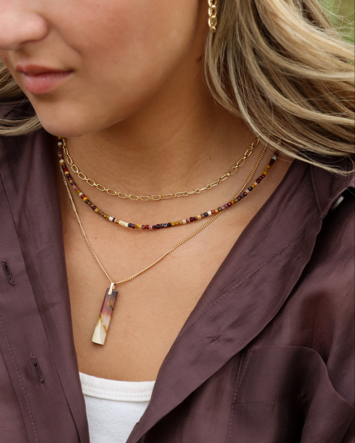 Woman wearing layered gold necklaces with a multicolor beaded strand and gemstone pendant, styled with a brown button-up shirt.