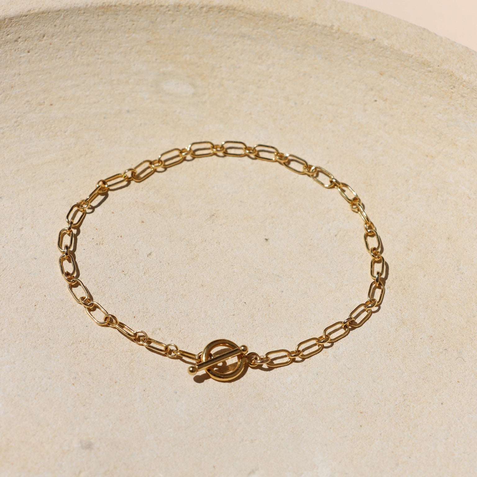 Gold paperclip-style link chain bracelet centered with a classic T-bar toggle clasp closure, displayed flat on a warm, textured stone tray in bright natural sunlight.