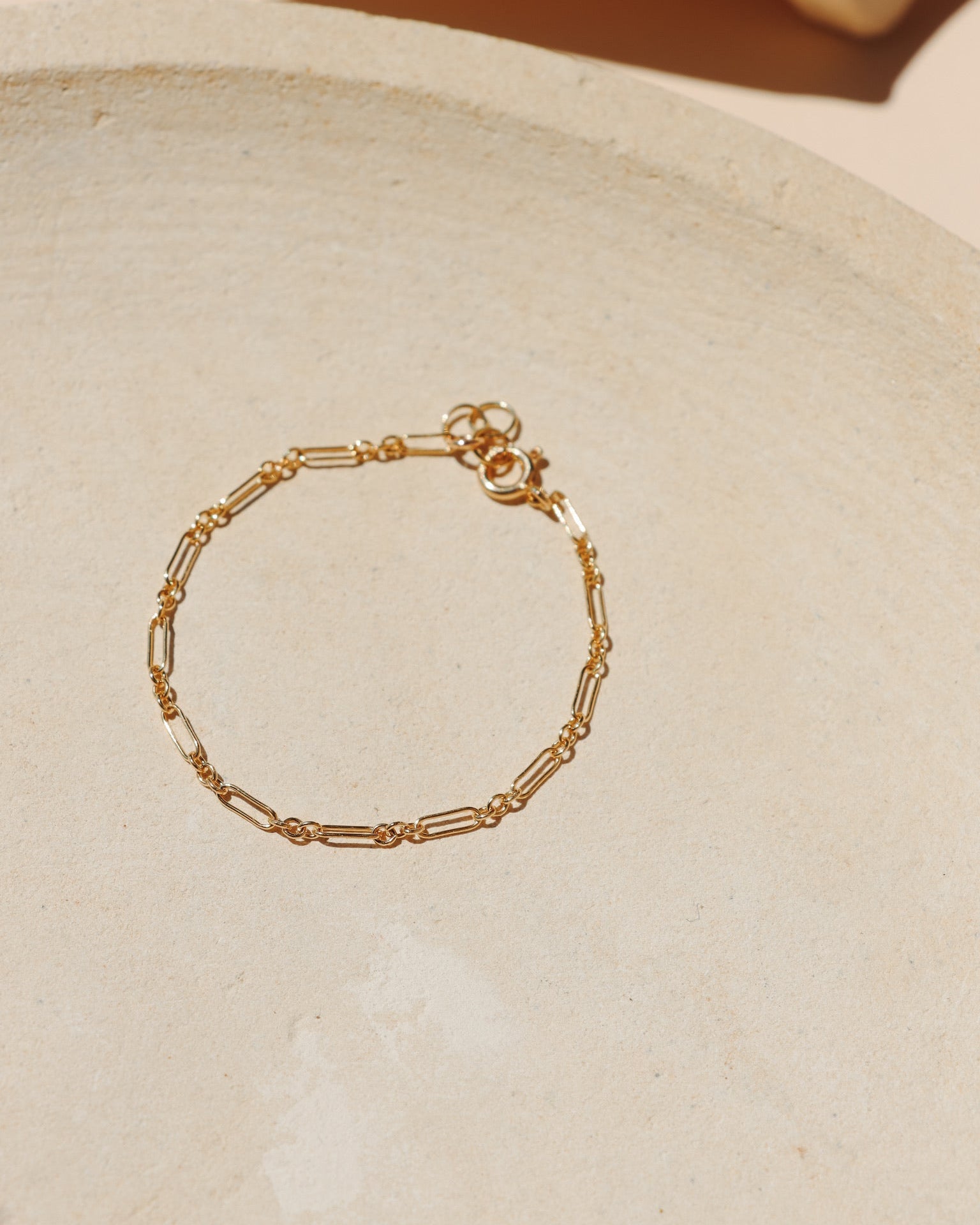 Minimalist gold chain bracelet with delicate elongated rectangular links and a spring ring clasp, arranged on a neutral, textured stone tray under soft natural lighting.