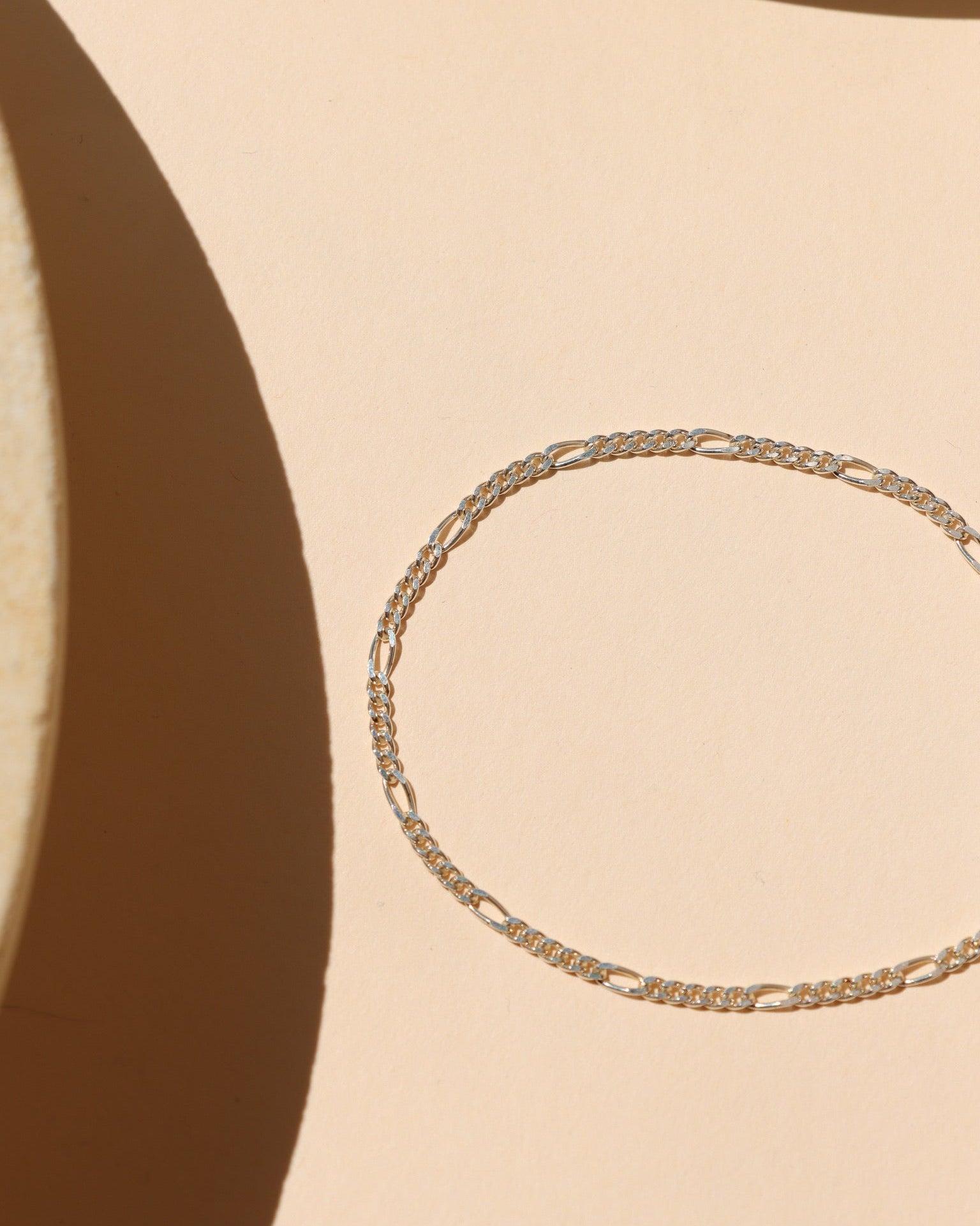 Minimalist sterling silver Figaro chain bracelet featuring alternating short round links and elongated oval links, laid flat on a peach-beige surface with geometric natural light shadows.