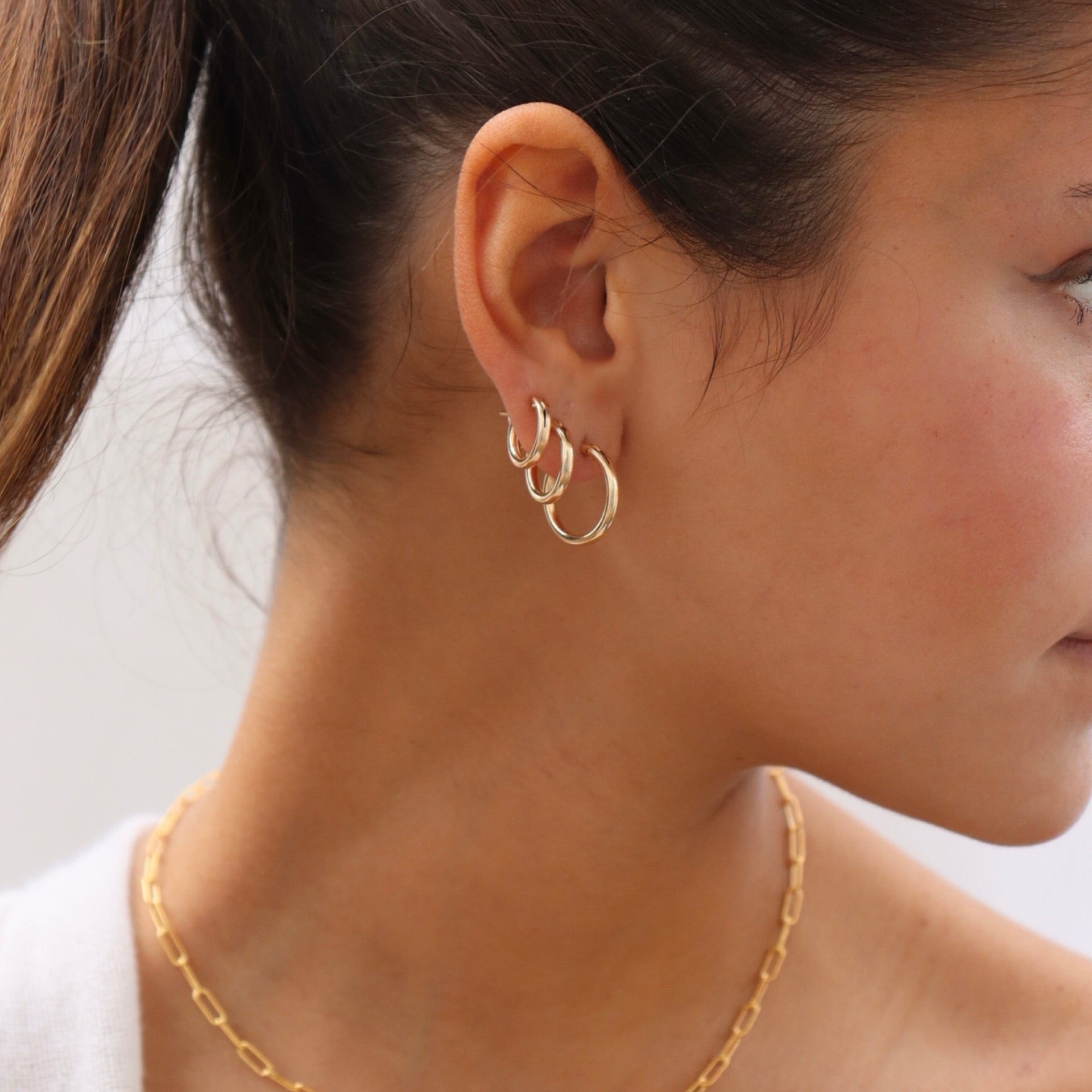 Side profile of a woman with brown hair tied in a ponytail, showcasing a ear stack of three polished gold hoop earrings graduated in size from smallest to largest along her lobe, paired with a gold paperclip chain necklace.
