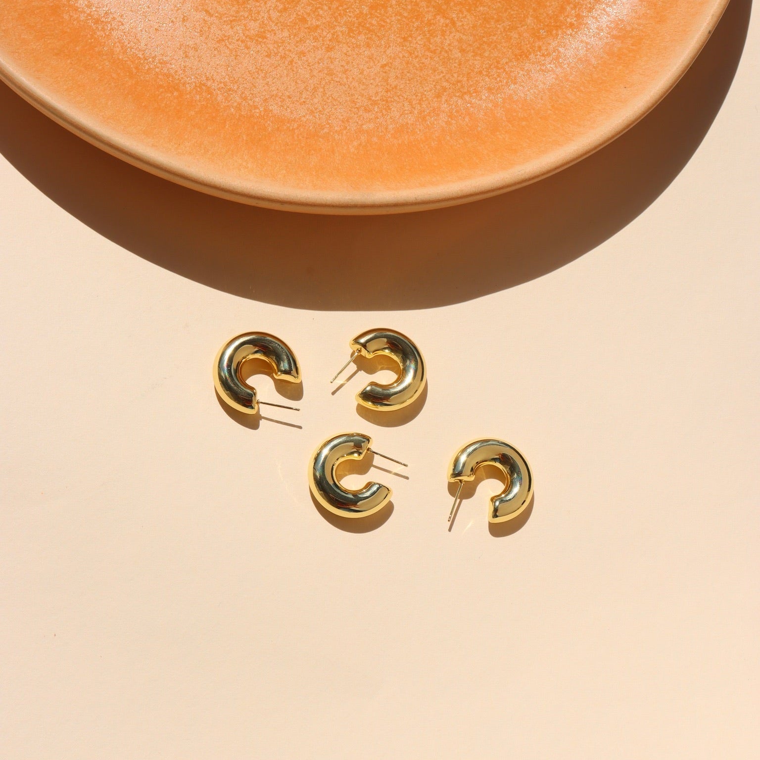 Top-down view of two pairs of thick, polished gold crescent hoop earrings placed on a light beige backdrop under bright, direct sunlight, with a warm terracotta-orange ceramic dish framing the top edge.
