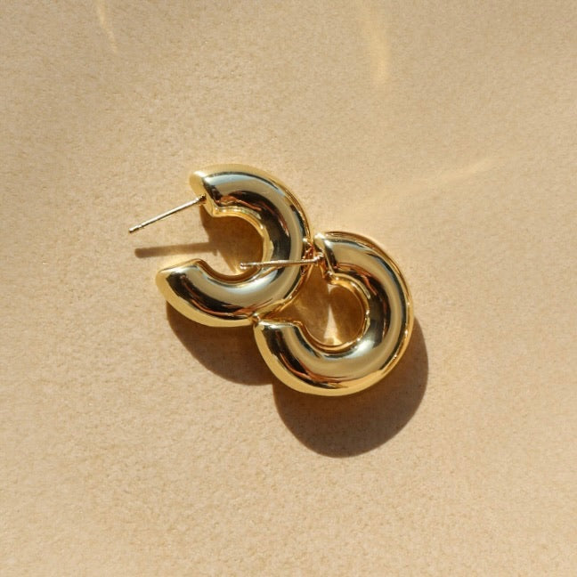 Top-down close-up view of a pair of chunky, hollow-style gold crescent hoop earrings overlapping each other on a textured beige surface, illuminated by warm sunlight with soft shadows