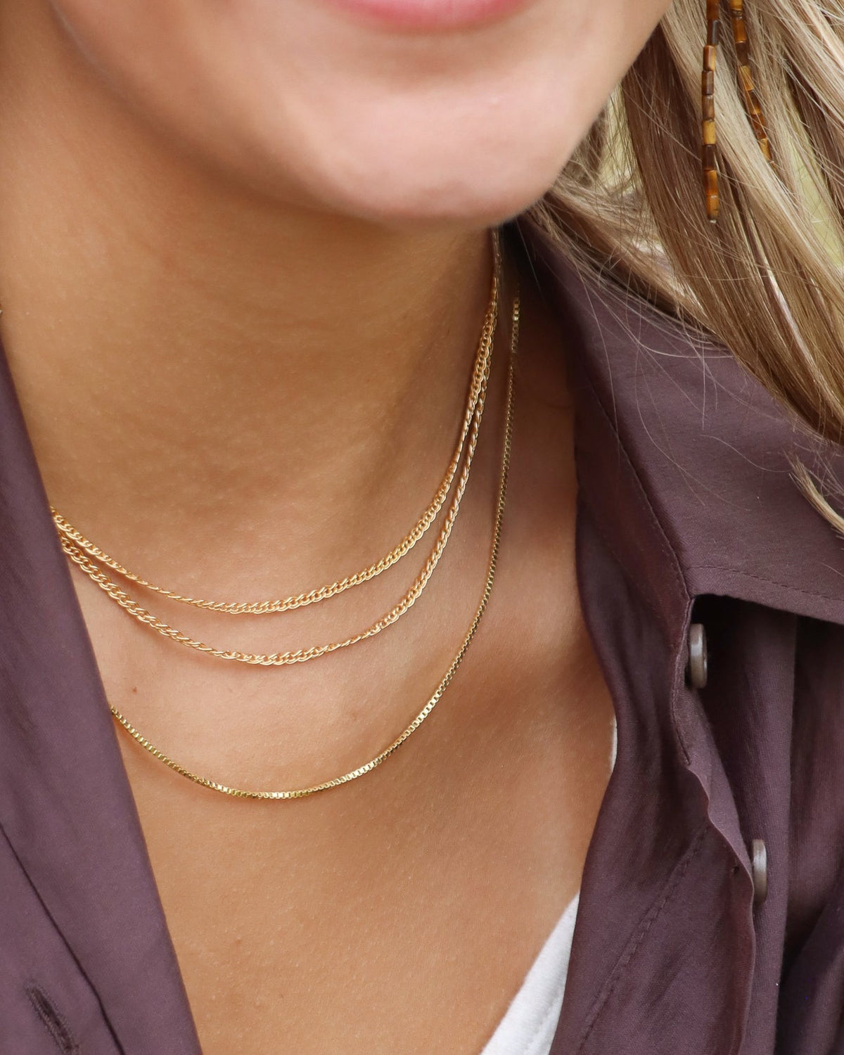 Woman wearing layered gold chain necklaces and long tiger’s eye earrings, styled with a brown button-up shirt.