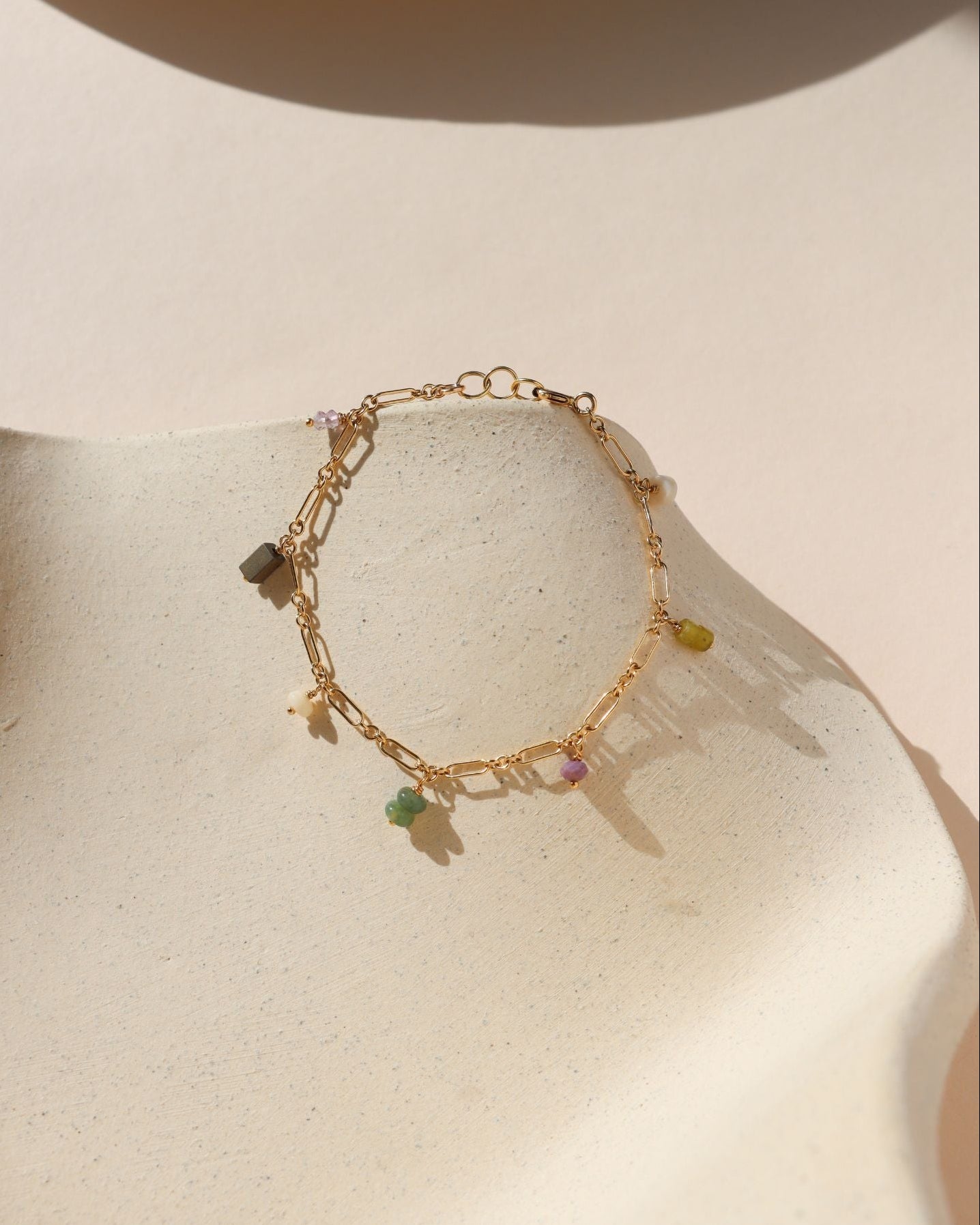 Gold chain linked bracelet with gemstone charms evenly spaced across it, laid on a beige dish in the sunlight