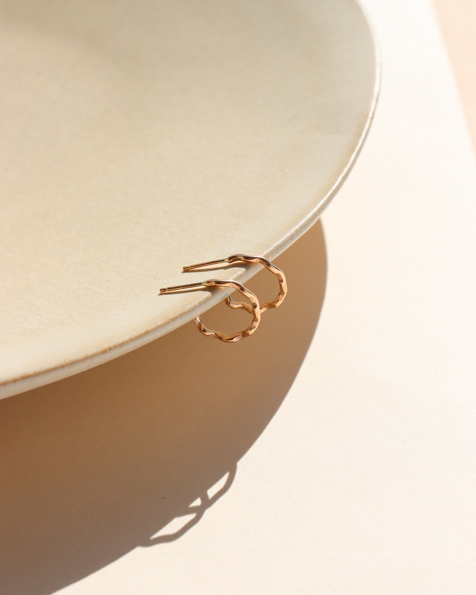 A wave like 14k gold fill textured huggie earring, displayed on a tan dish in the sunlight. 