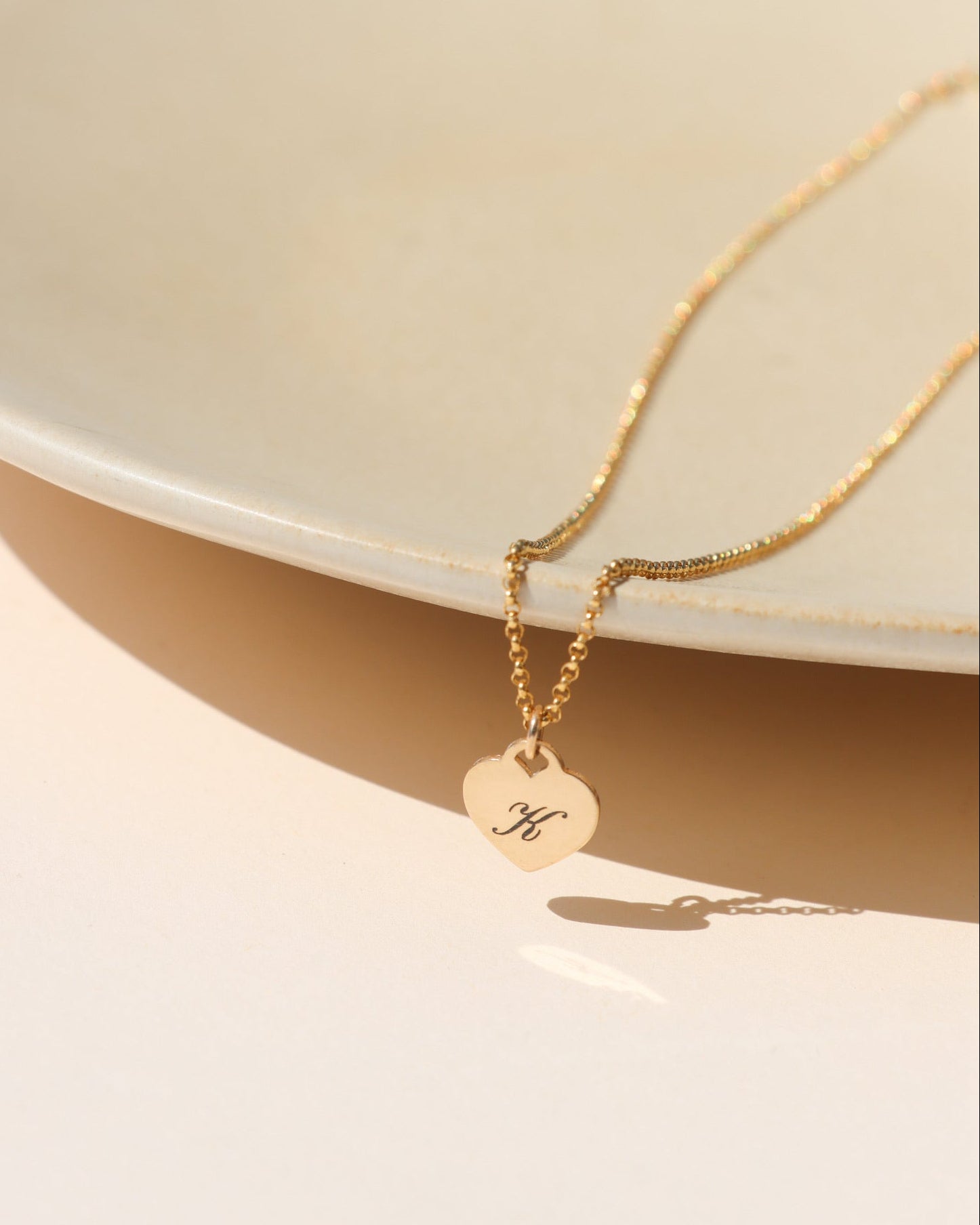 Gold heart pendant necklace engraved with the letter K, styled on a ceramic dish in warm sunlight.