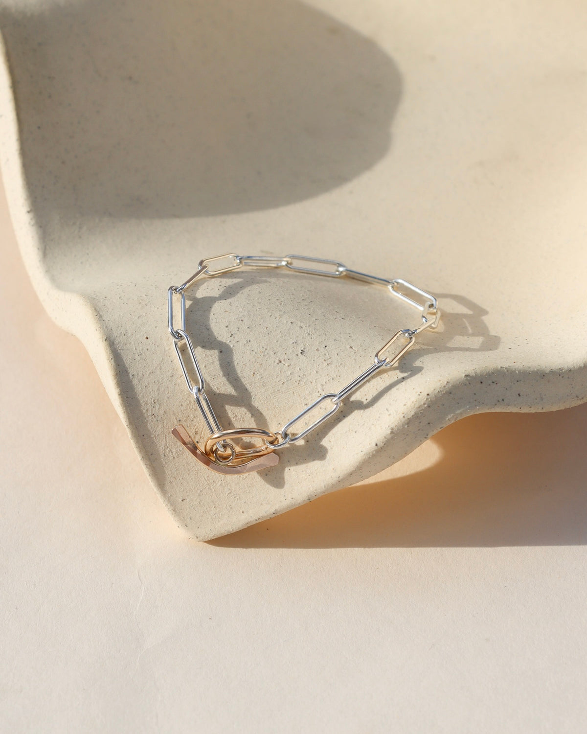 Flux Toggle Bracelet featuring elongated sterling silver paperclip chain links and a gold fill toggle clasp, displayed on a textured cream ceramic surface in natural sunlight.