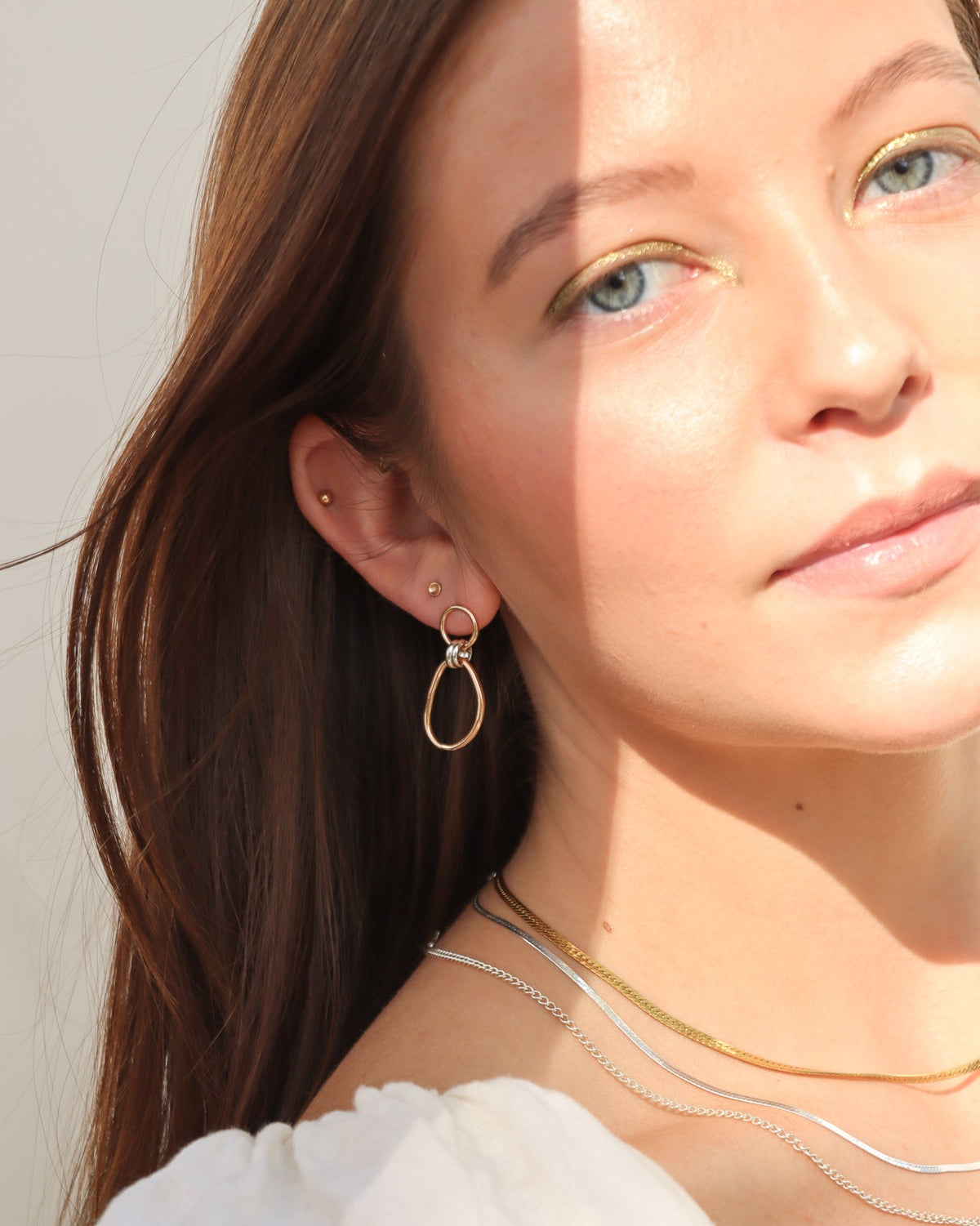 Close-up of a model wearing mixed-metal drop earrings with gold oval hoops and silver connecting rings, styled with layered gold and silver necklaces in natural sunlight.
