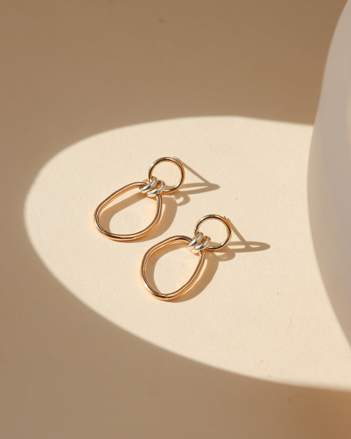 Mixed-metal drop earrings featuring small gold circular studs, silver connecting rings, and elongated gold oval hoops displayed on a cream background in warm sunlight.