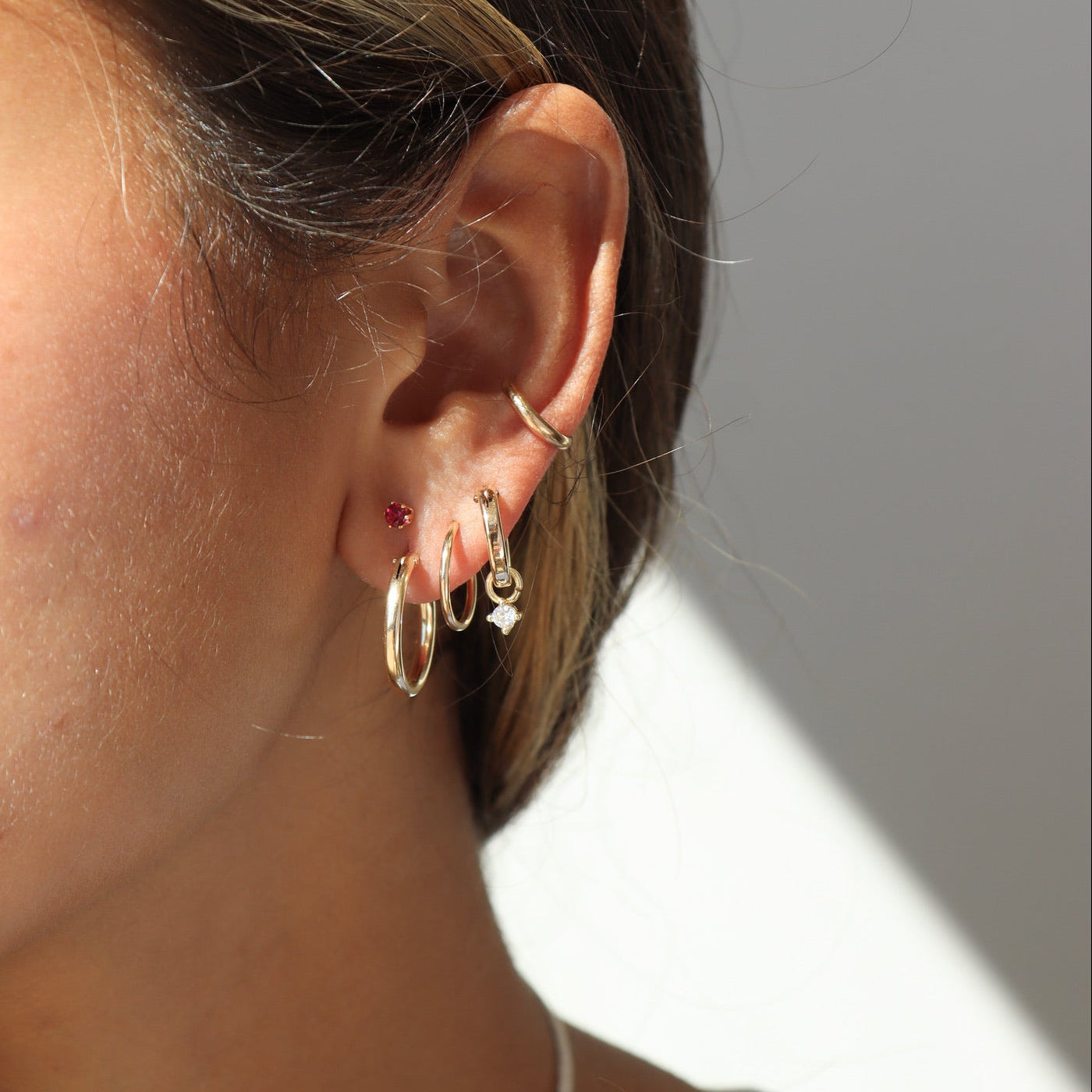 Side view of an ear stack featuring three gold lobe hoops—including one with a sparkling diamond dangle charm—a tiny red gemstone stud, and a minimal smooth gold ear cuff along the mid-cartilage.