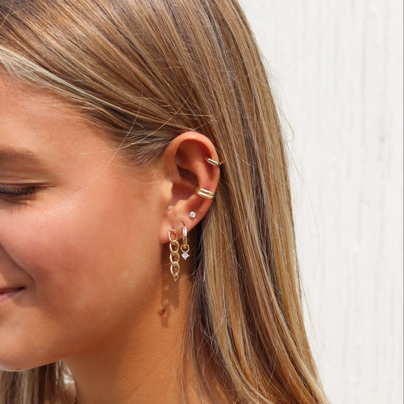Woman wearing layered gold necklaces, gold hoop and chain earrings, a diamond stud, and gold ear cuffs.