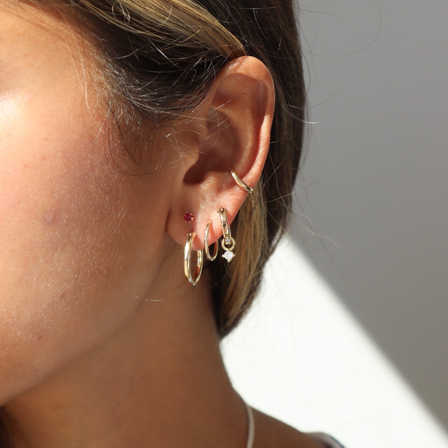 Side view of an ear stack featuring three gold lobe hoops—including one with a sparkling diamond dangle charm—a tiny red gemstone stud, and a minimal smooth gold ear cuff along the mid-cartilage.
