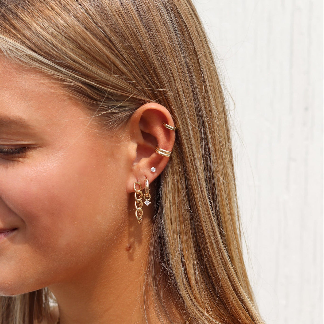 Woman wearing layered gold necklaces, gold hoop and chain earrings, a diamond stud, and gold ear cuffs.
