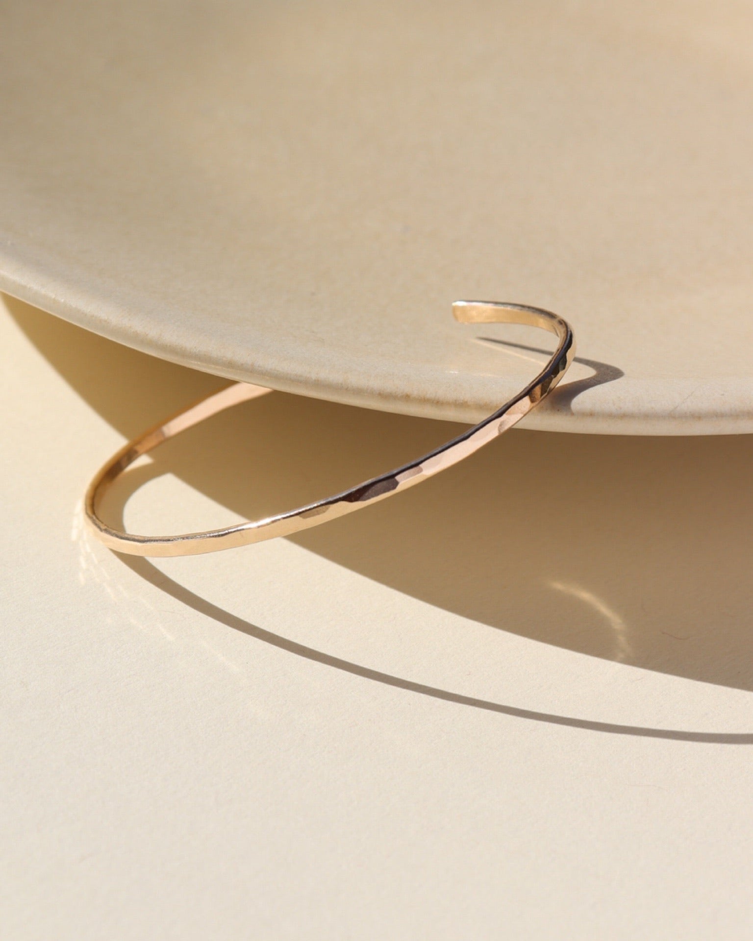 14k gold filled cuff with lightly hammered texture leaned on a beige ceramic dish in the sunligjt. 