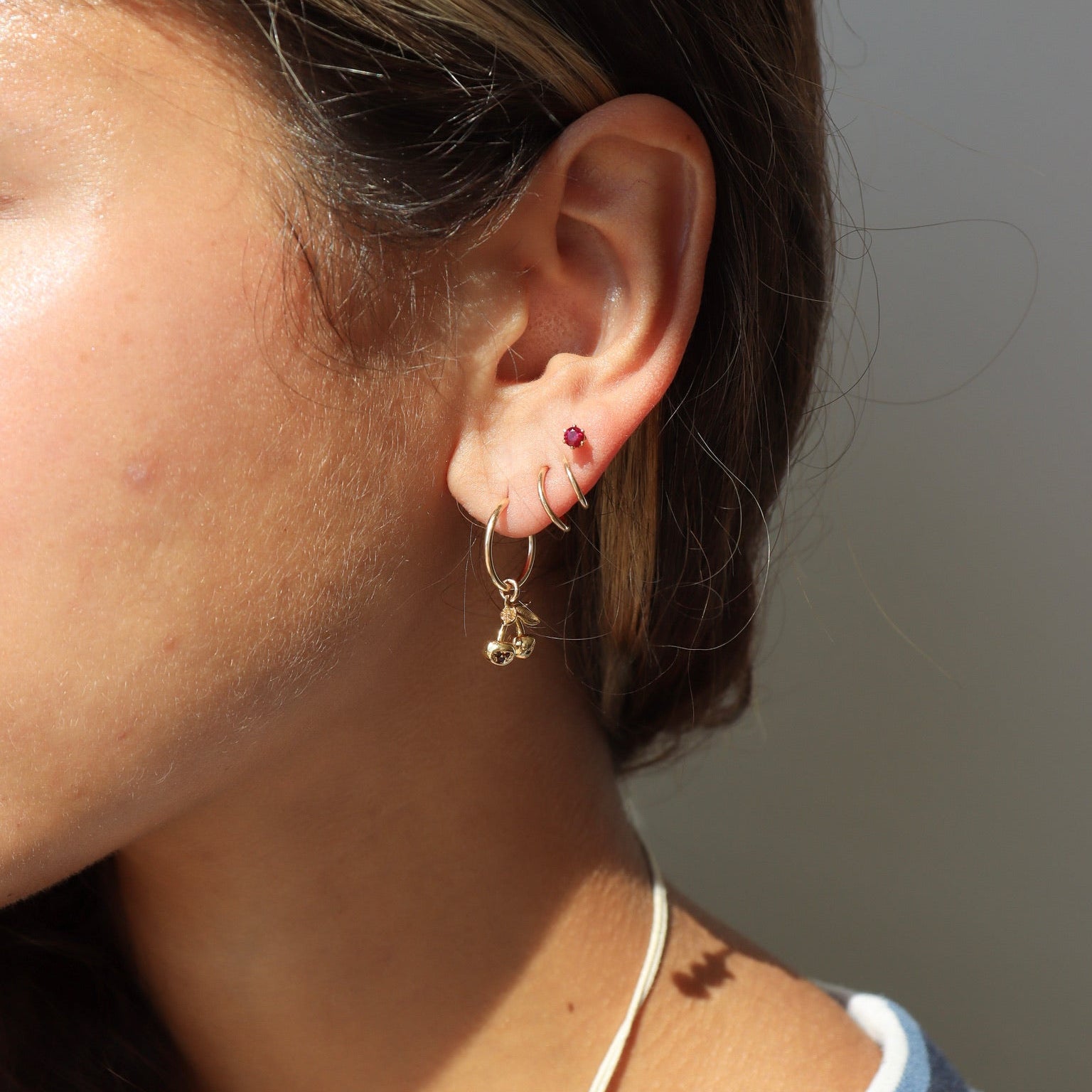 Side profile shot of a woman with her hair pulled back, showcasing a four-piercing ear stack in natural sunlight: a small gold hoop with a dangling cherry charm in her lobe, two thin gold huggie hoops in her second and third placements, and a tiny red gemstone stud.