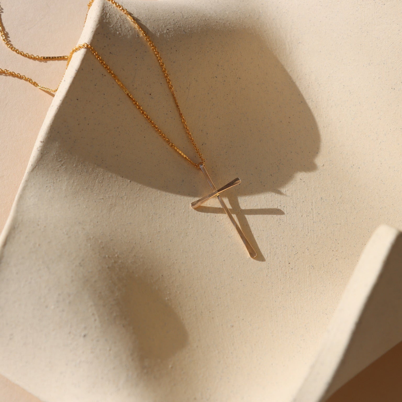 Grace Cross Necklace in 14k gold fill featuring a modern cross pendant on a delicate chain displayed on a textured ceramic surface.