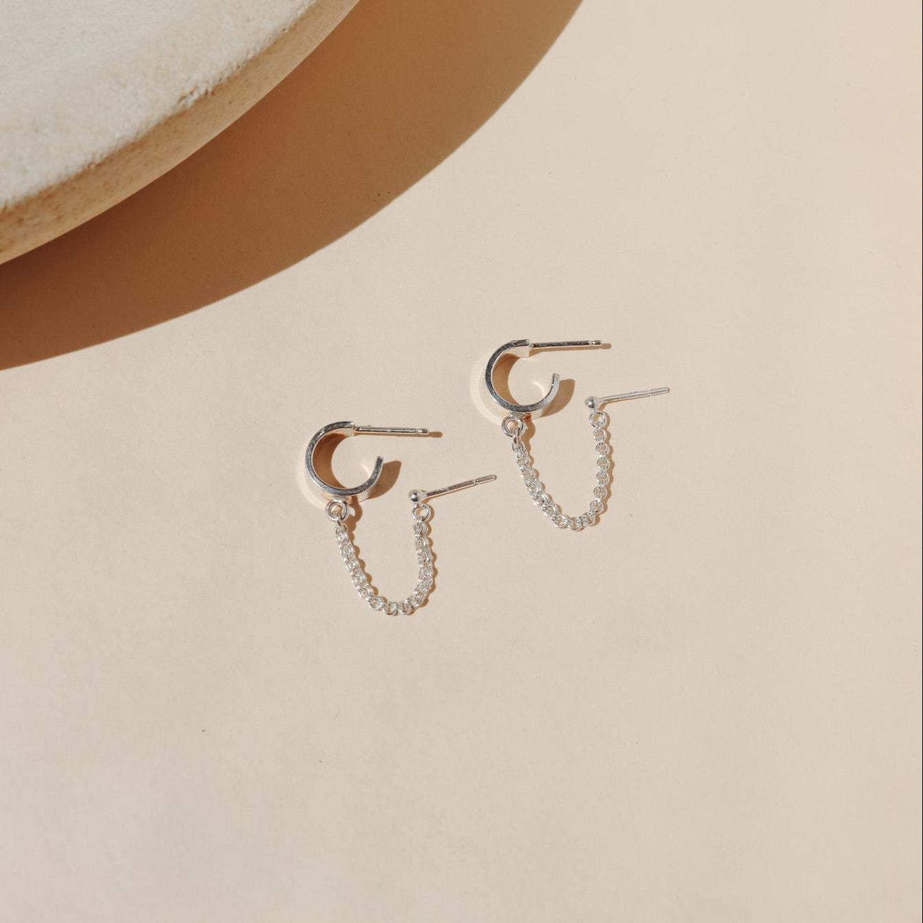 A sterling sliver hoop is connected to a sterling silver classic stud earring by a rolo silver chain on a beige background in the sunlight