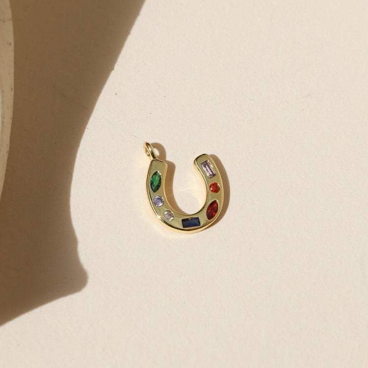 Gold horseshoe shaped charm with green, clear, blue, and red CZ gems embedded within, laid on a beige background in the natural sunlight. 