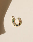 Gold horseshoe shaped charm with green, clear, blue, and red CZ gems embedded within, laid on a beige background in the natural sunlight. 